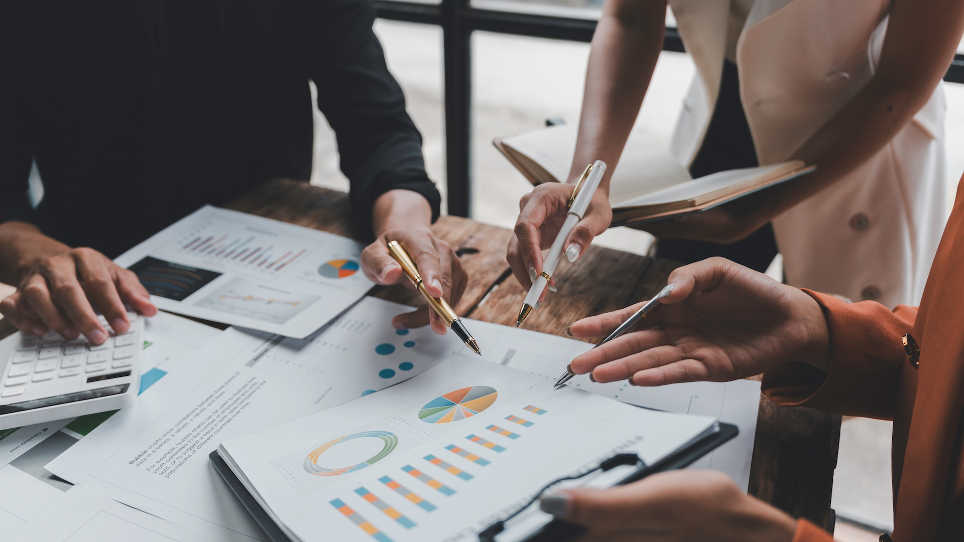 Close-up of a group of professionals collaborating around a wooden table, analyzing financial charts and graphs on printed documents. One person is using a calculator, while others are pointing at reports with pens, discussing data and strategy.